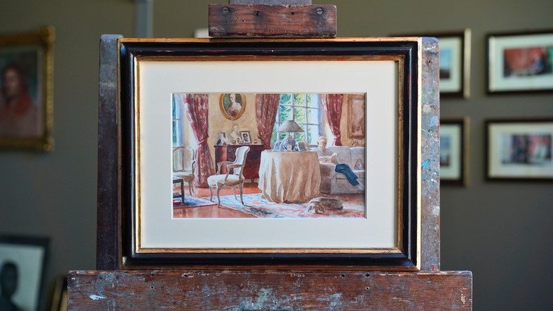 A framed interior watercolour standing on a wooden easel: a red-curtained room with a round table and chairs