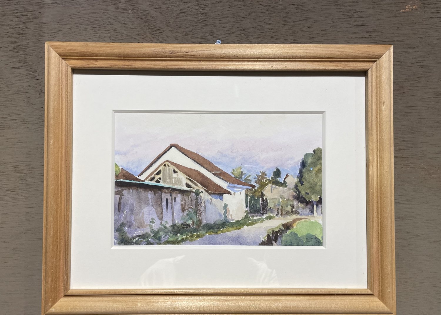 A framed watercolour hanging on a grey wall: a pale natural wood moulding, a deep white bevelled mount, and behind glass a painting of a tiled Balinese house beside a lane